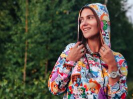 Vintage Graphic Hoodie: Retro Street Style Guide A young woman stands outdoors in a colorful hooded sweatshirt covered with cartoon characters. She’s turned to the side, looking away from the camera, with a forest backdrop. She wears rings, a necklace, and a large watch.