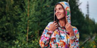 Vintage Graphic Hoodie: Retro Street Style Guide A young woman stands outdoors in a colorful hooded sweatshirt covered with cartoon characters. She’s turned to the side, looking away from the camera, with a forest backdrop. She wears rings, a necklace, and a large watch.