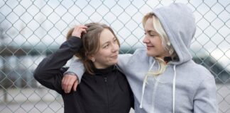 Vintage Zip Hoodie: Classic Retro Streetwear Style Two young women stand close together with their arms around each other in front of a chain-link fence. One has brown hair and wears a black hooded jacket; the other has blonde hair and wears a gray hoodie. They gaze at each other with warm smiles.