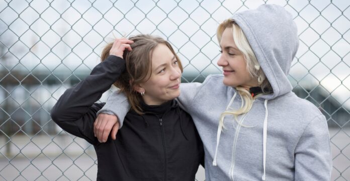 Two young women stand close together with their arms around each other in front of a chain-link fence. One has brown hair and wears a black hooded jacket; the other has blonde hair and wears a gray hoodie. They gaze at each other with warm smiles.
