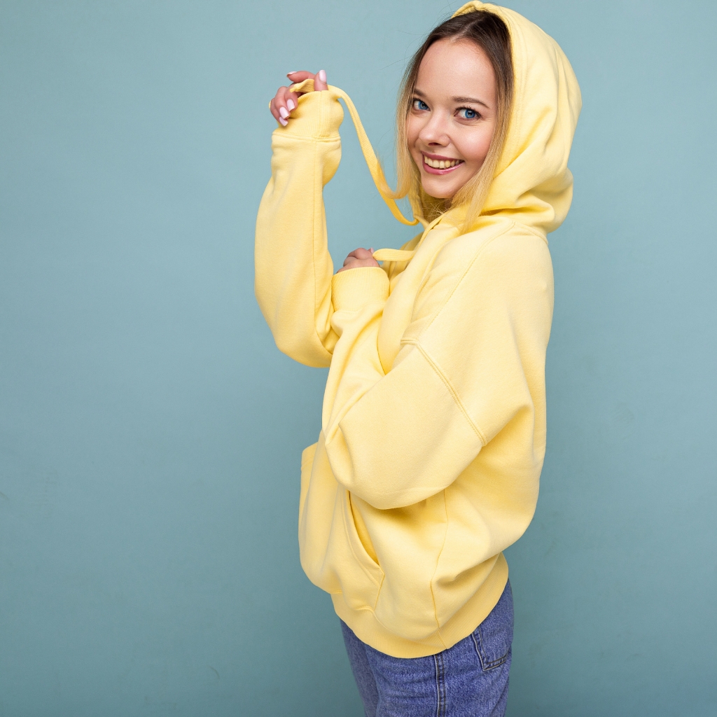 A smiling young woman with blue eyes and blonde hair wears a bright yellow hooded sweatshirt with the hood up, holding a drawstring. She stands against a light blue background.
