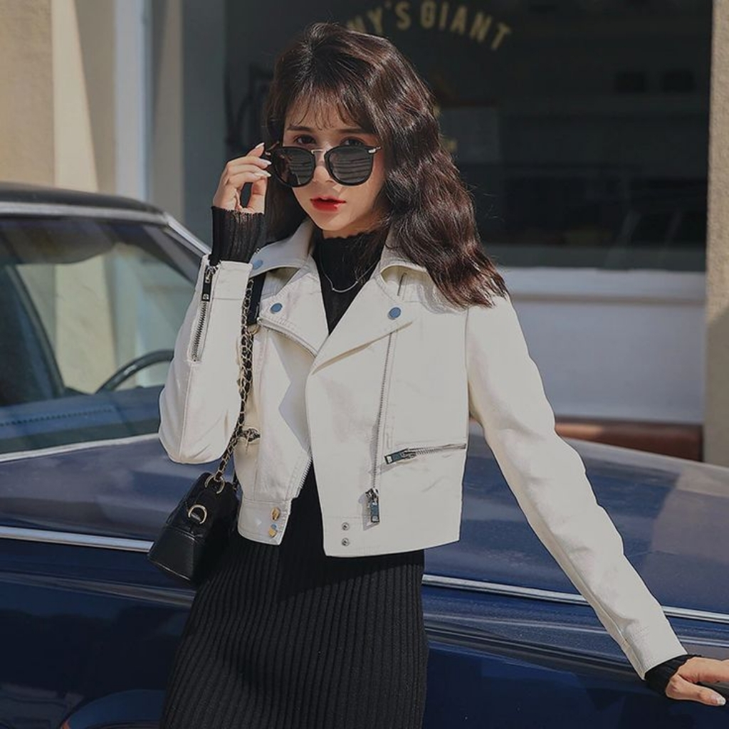 White Leather Jacket with Clean Minimalist Design 2 A woman with long wavy brown hair wearing a white biker jacket and black outfit stands beside a blue car outdoors. She holds black sunglasses near her face, with a small black shoulder purse. The scene is brightly lit, with a building and dark windows in the background.