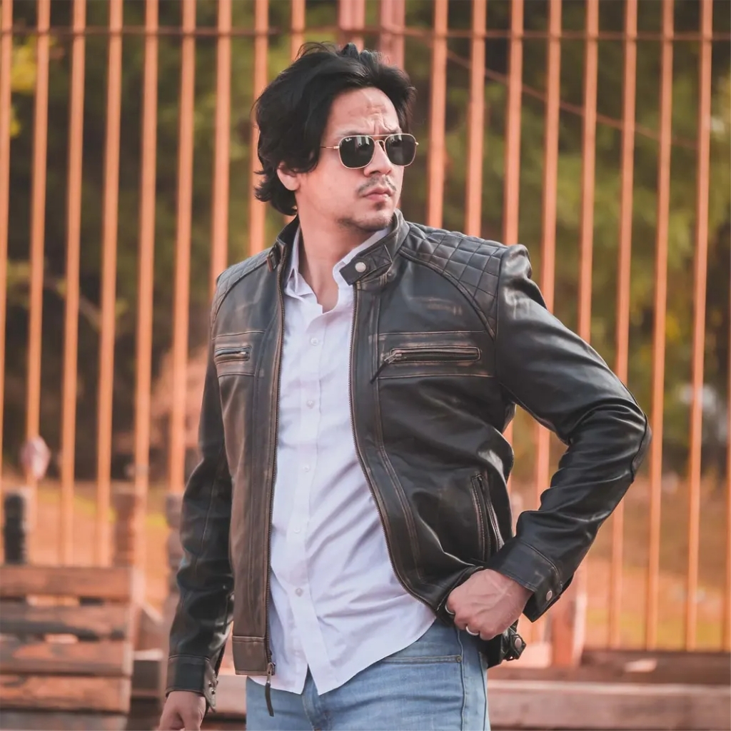 Premium Shirt Style Leather Jacket for Casual and Formal Looks 5 A man wearing a black leather jacket and sunglasses stands in front of a brown metal gate. He has dark hair and is wearing a white shirt and blue jeans, with his left hand in his pocket.