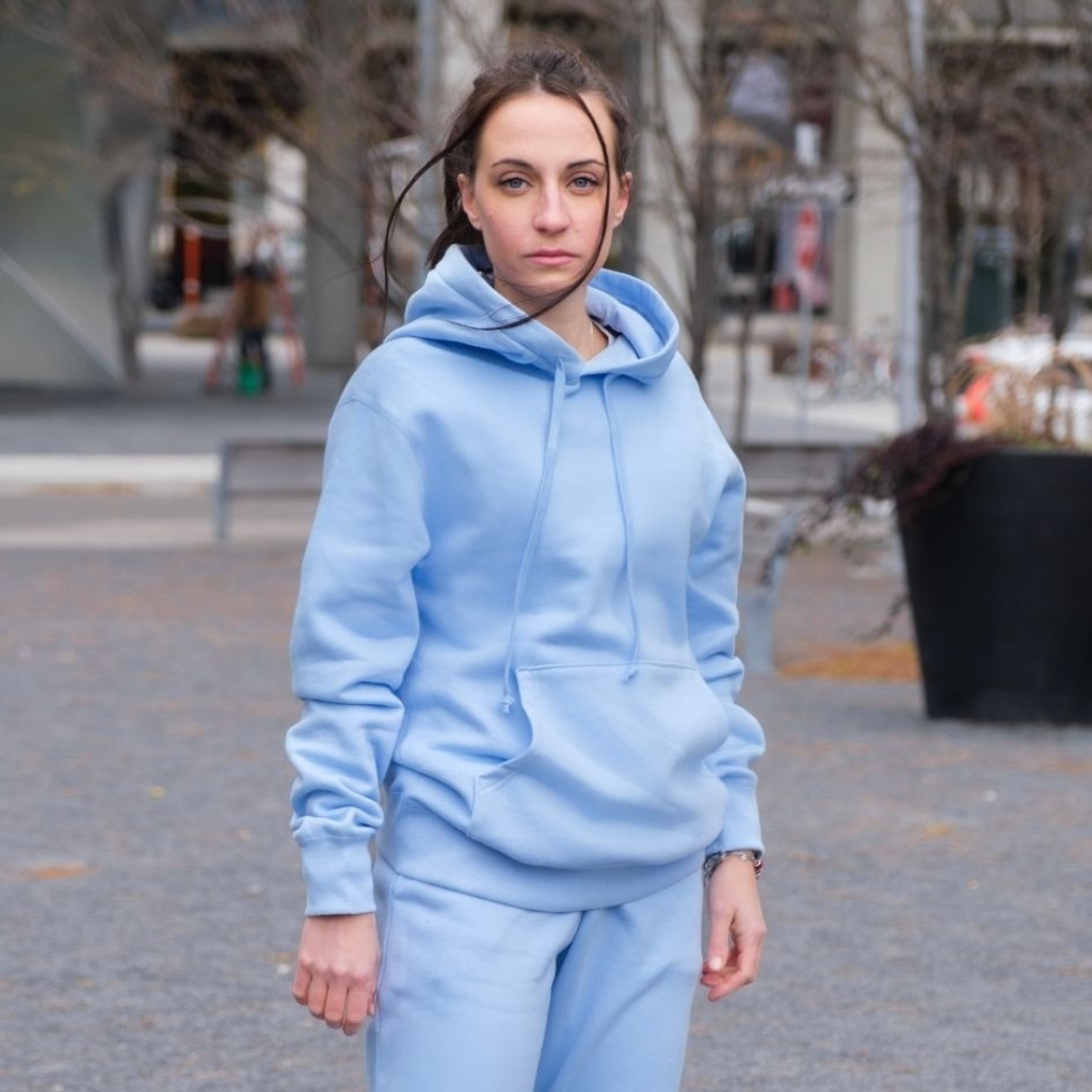 Logo Graphic Hoodie: Trendy Streetwear Style Guide 6 A woman stands outdoors in a light blue matching hoodie and sweatpants set, facing the camera with a neutral expression. The background shows a plaza with benches, trees, and buildings on an overcast day.