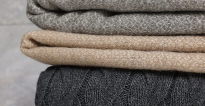Woven Pure Cashmere Fabric Close-up of three folded knit blankets stacked in dark gray cable knit, light beige textured weave, and gray diamond-pattern knit, with a soft, cozy appearance and a blurred light gray background.