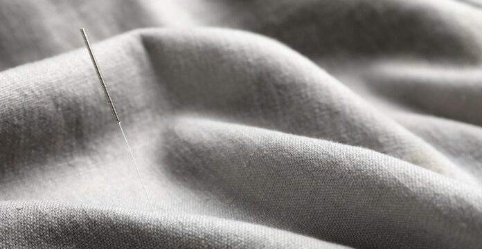 Woven Recycled Cashmere Fabric An acupuncture needle inserted into gray woven fabric, showing a thin silver needle with a textured handle amid softly draped folds.