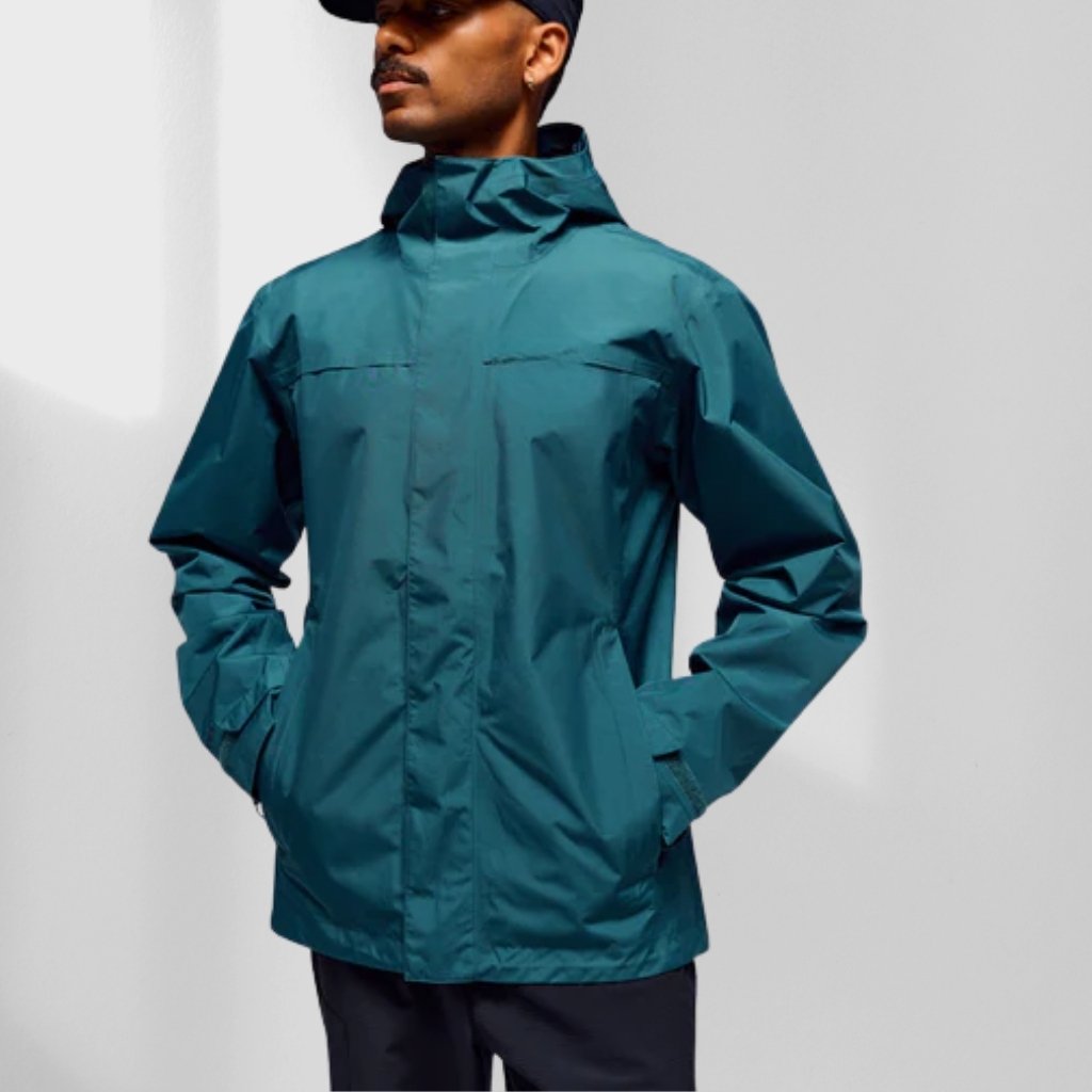 A person wearing a teal hooded rain jacket with their hands in their pockets, standing against a white background. They are wearing a dark baseball cap.