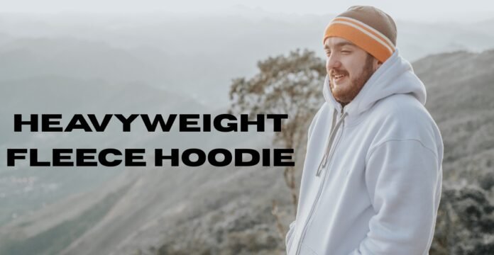 A man with a beard wearing an orange-and-white striped beanie and a light-colored fleece hoodie stands outdoors on a misty mountain overlook.