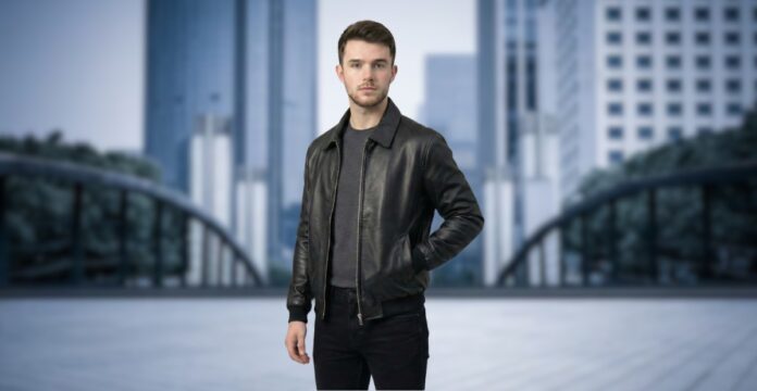 A man standing outdoors in an urban setting, wearing a black nubuck leather jacket, a dark gray sweater, and black jeans. The cityscape in the background is blurred.