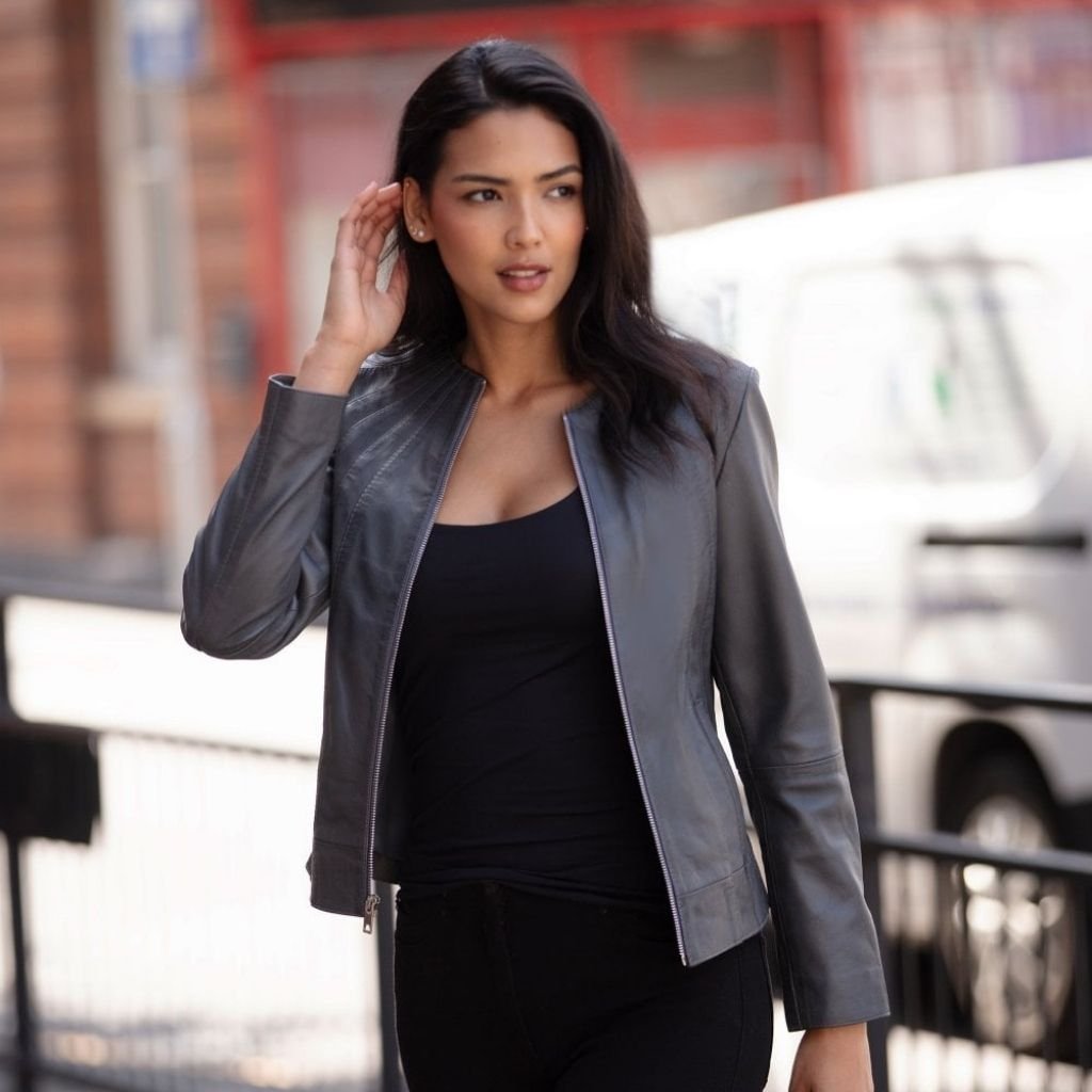 Grey Leather Jacket – Stylish & Versatile Outerwear for Every Look 6 A woman on a city street wearing a gray leather jacket over a black top and black pants. She has dark hair and is touching her right ear while looking off to the side. Background shows a brick building with red trim, a white van, and a black metal fence.