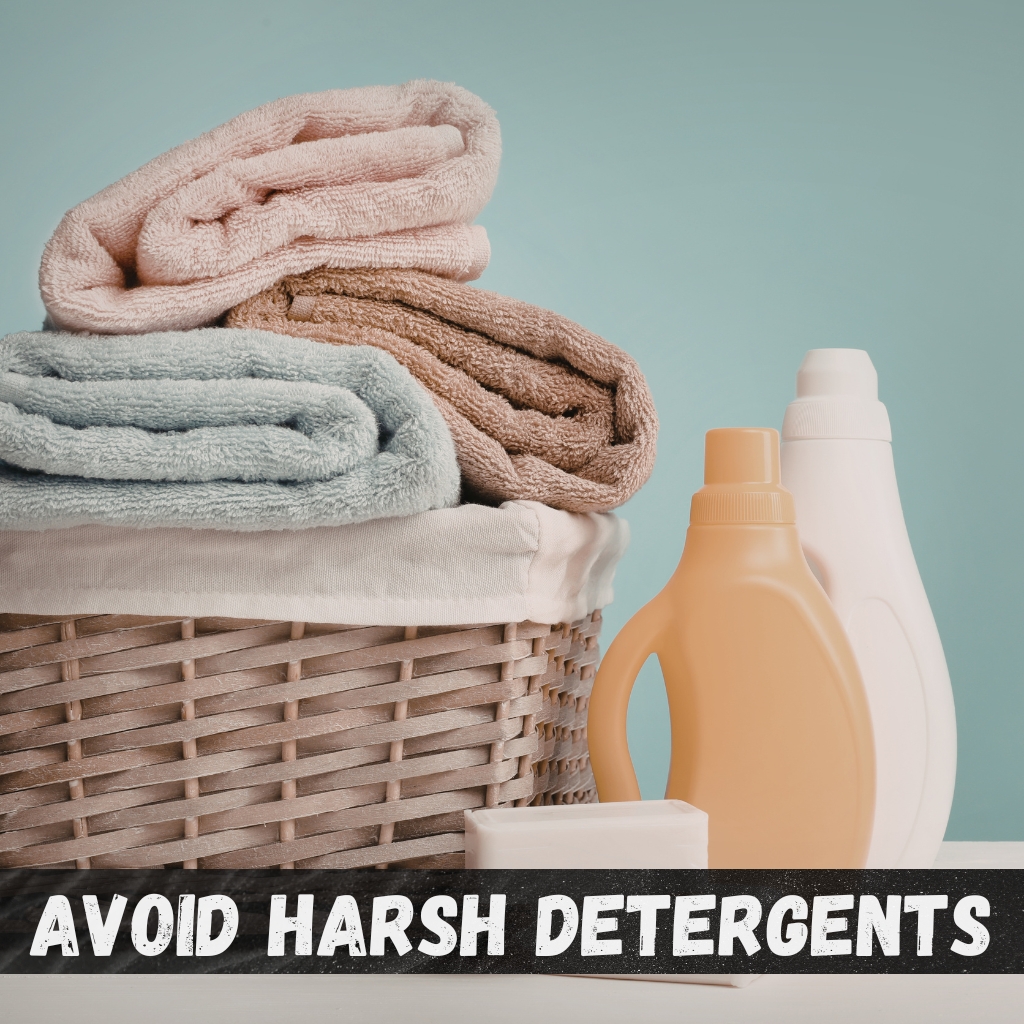 A wicker basket filled with neatly folded towels in light blue, beige, and pink. Beside the basket are two detergent bottles (one white, one light orange) and a bar of soap. The background is a soft blue, with a black banner at the bottom that reads “AVOID HARSH DETERGENTS.”