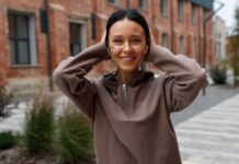 Cropped Half-Zip Hoodie: Stylish Comfort for Modern Fashion A smiling woman with dark hair wearing a brown fleece hoodie, touching her ears, standing on a sidewalk with brick buildings and trees in the background.