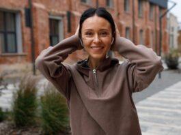A smiling woman with dark hair wearing a brown fleece hoodie, touching her ears, standing on a sidewalk with brick buildings and trees in the background.