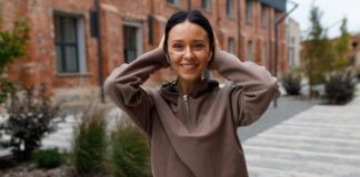 Cropped Half-Zip Hoodie: Stylish Comfort for Modern Fashion A smiling woman with dark hair wearing a brown fleece hoodie, touching her ears, standing on a sidewalk with brick buildings and trees in the background.