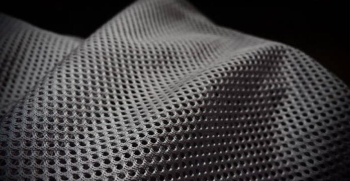 Close-up of a gray, net-like fabric with a grid of small, evenly spaced holes and subtle folds. The background is dark and blMaximum Warmth Winter Coat fabric texture: close-up of a gray mesh-like material. urry.Air Mesh Pro Performance Fabric: