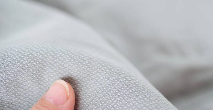 Alt text: A close-up of light gray fabric with a subtle repeating pattern, slightly bunched, with a finger touching the fabric in the lower left corner. Chill Tech Moisture Control Fabric: