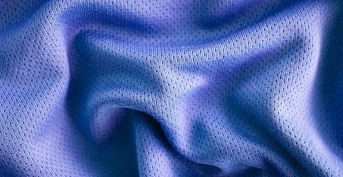 A close-up photo of a textured, blue fabric with a smooth, crumpled surface, showing small perforations or mesh-like holes. Vapor Flow Athletic Fabric: