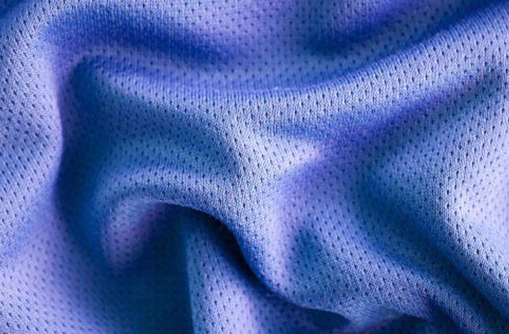 A close-up photo of a textured, blue fabric with a smooth, crumpled surface, showing small perforations or mesh-like holes. Vapor Flow Athletic Fabric: