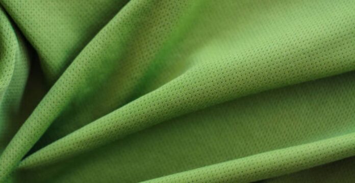 A close-up photograph of a bright green, textured fabric with small perforations. The fabric is folded in soft, overlapping layers, creating smooth curves and subtle shadows that emphasize its breathable, mesh-like weave.
