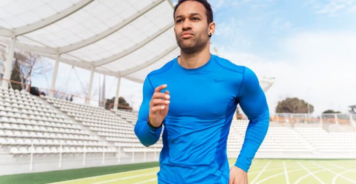 A man in a bright blue athletic long-sleeve shirt runs on a track in a stadium with empty white seats and a clear sky in the background. Power Fit Compression Fabric: