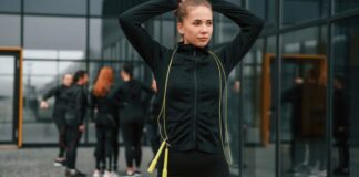 Gym and Fitness Training Outfits: Style, Comfort & Results A young woman in athletic wear stands outdoors with her hands lifting her hair, preparing for exercise. She has a yellow jump rope around her neck. In the background, a group of athletes stands near a modern building with large glass windows and orange-framed doors. Gym and Fitness Training Outfits: