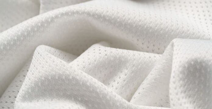 Close-up of white, textured fabric with tiny perforations arranged in a subtle repeating pattern, draped in soft folds to show light and shadow.Flex Pro Sports Fabric: