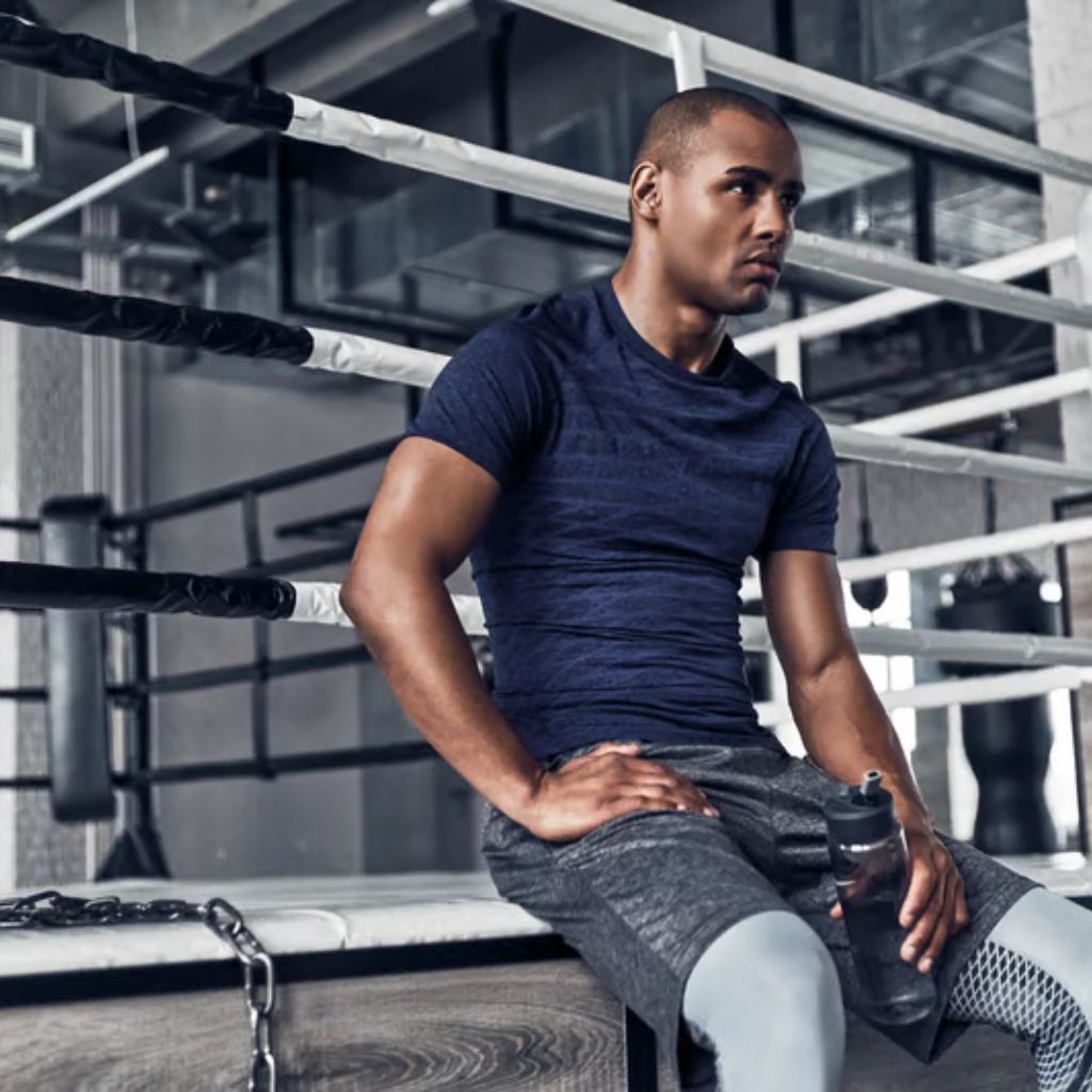A man sits on the edge of a boxing ring, wearing a blue t-shirt, grey shorts, and compression leggings. He holds a water bottle and looks to the side. The boxing ring is black and white with a grey wall in the background.
Gym and Fitness Training Outfits: