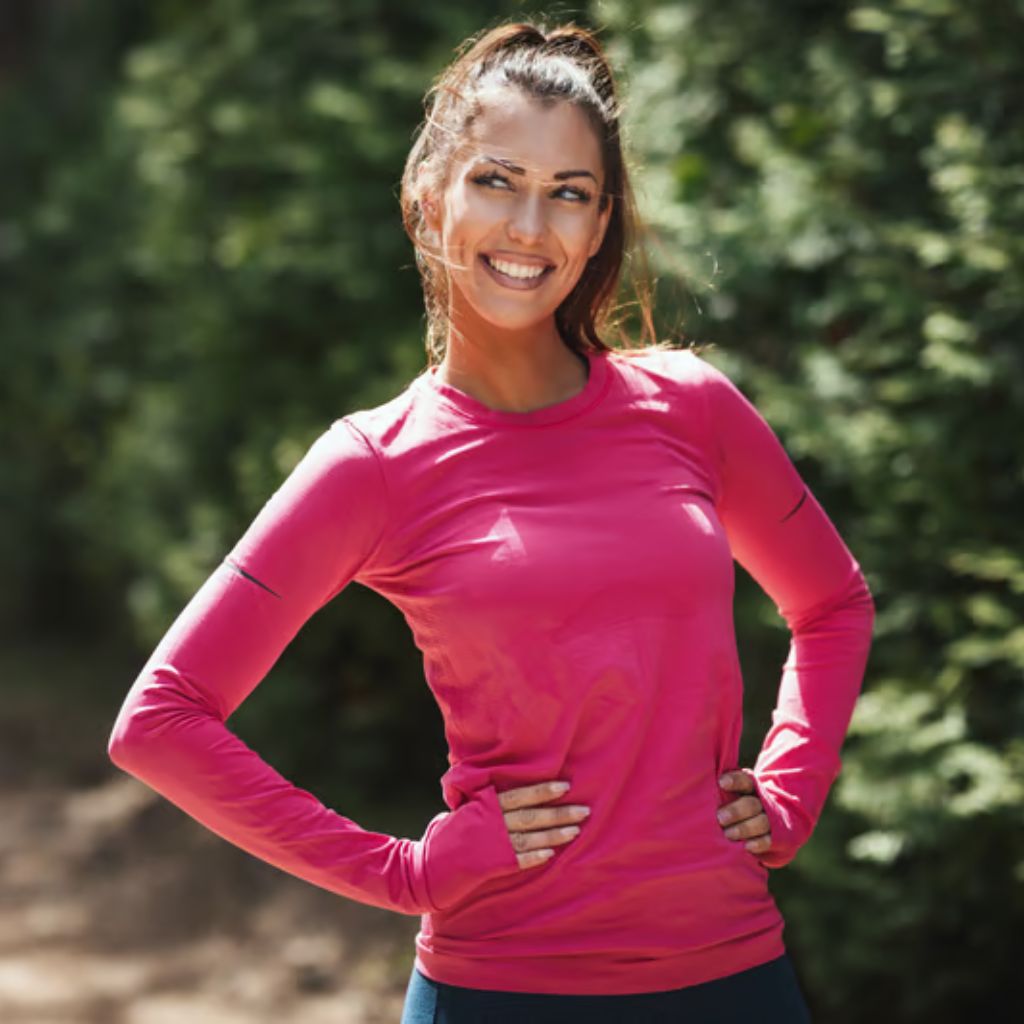 A young woman with brown hair tied back stands outdoors with hands on her hips, wearing a long-sleeved pink top and blue leggings. The background shows blurred green trees.
Sports Shirts and Athletic Tops: