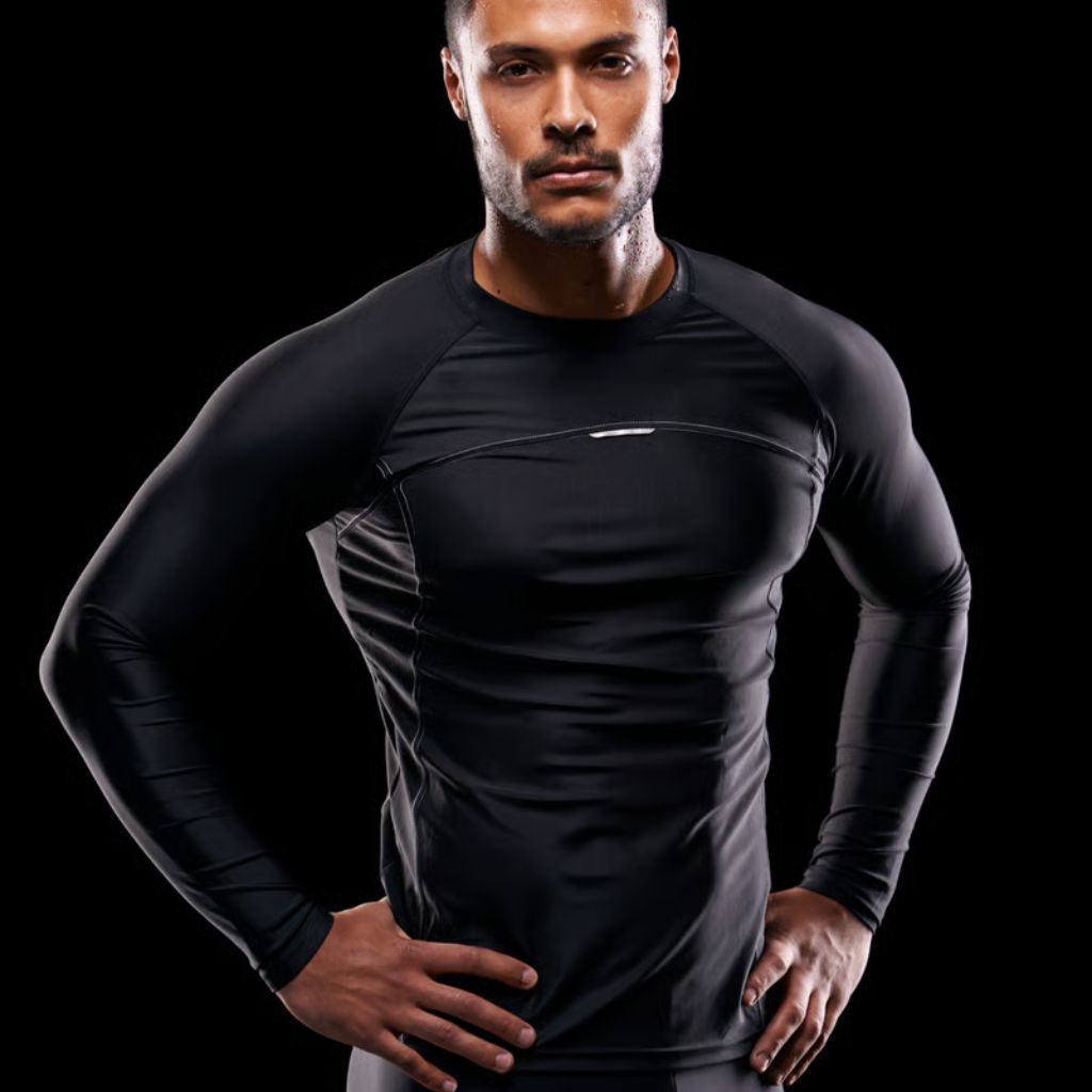 A muscular man with short dark hair and a neatly trimmed beard stands against a solid black background. He wears a tight-fitting black long-sleeve shirt and dark pants or shorts, with his hands on his hips, looking directly at the camera with a serious expression.
Power Fit Compression Fabric: