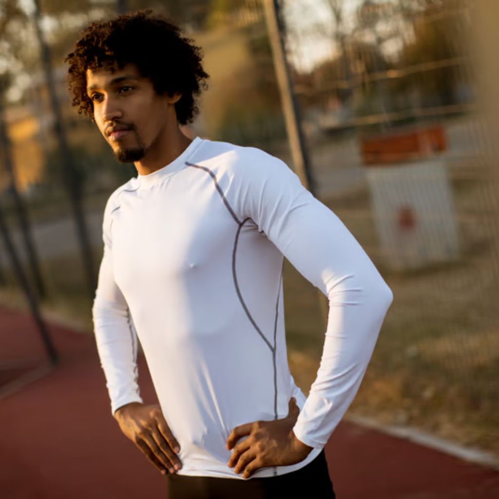 A young Black man with curly hair stands outdoors with hands on hips, wearing a white long-sleeved athletic top and black leggings. The background shows a blurred fence and trees.Sports Shirts and Athletic Tops:
