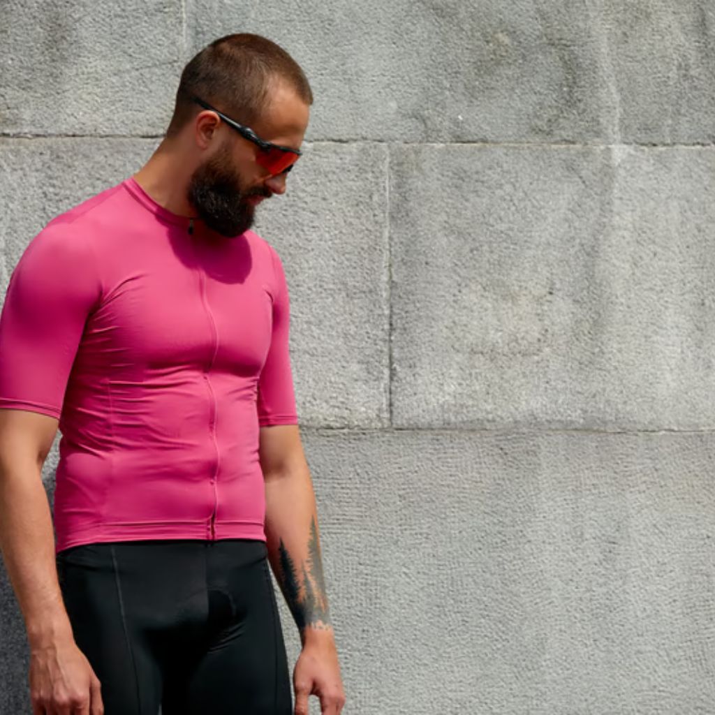 A bearded man wearing a bright pink athletic cycling jersey with a zipper and black cycling shorts stands against a textured gray concrete wall. He has red-tinted sunglasses and is looking downward; a dark green tree tattoo is visible on his left forearm.
Power Fit Compression Fabric: