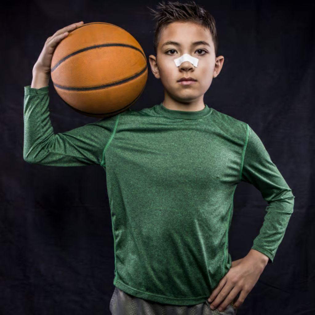 A young boy stands against a dark background, wearing a green long-sleeved shirt and gray shorts. He holds a basketball on his shoulder with his right hand and rests his left hand on his hip. He has a white bandage on his nose.
Sports Shirts and Athletic Tops:
