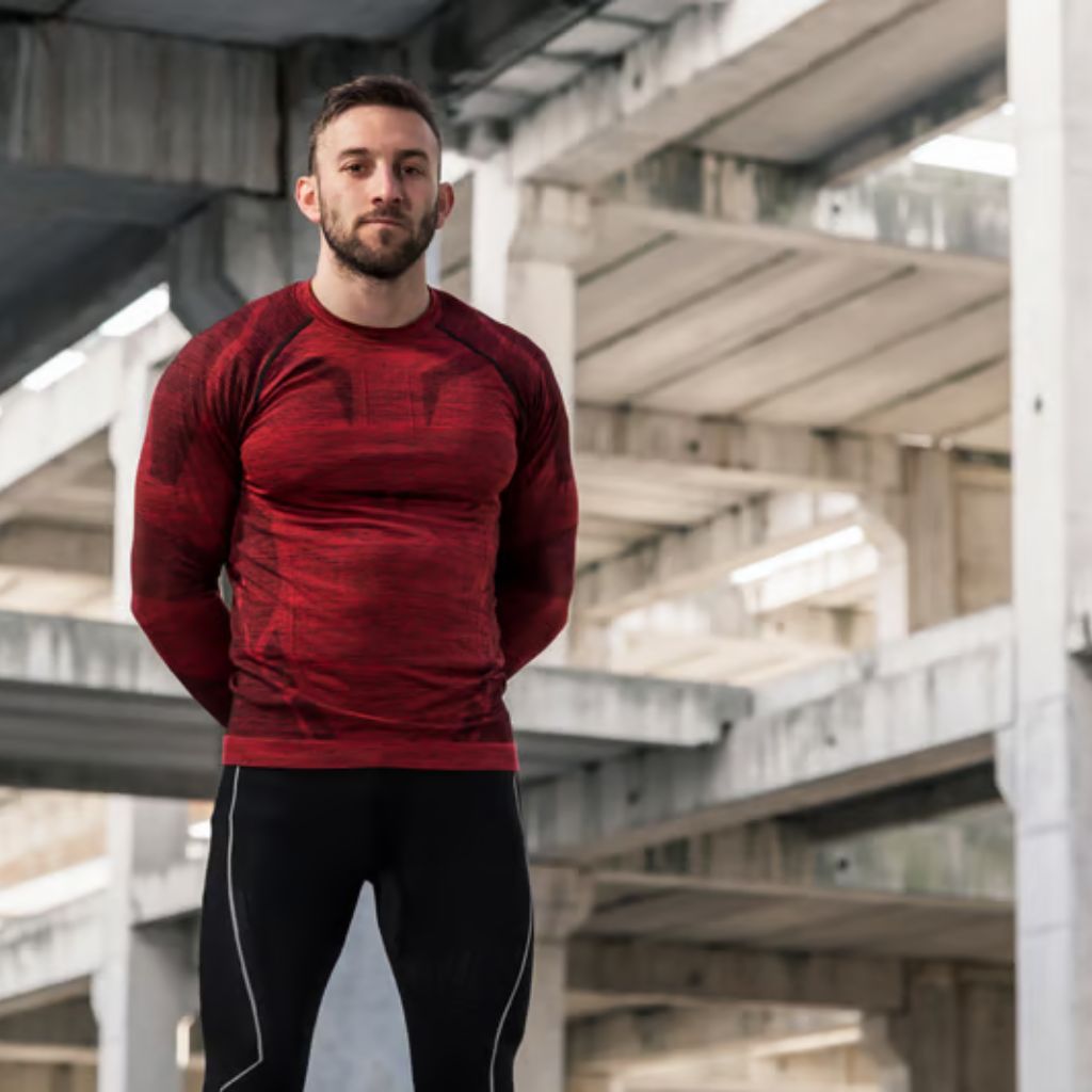 A bearded man stands outdoors in front of a concrete structure with multiple beams. He wears a long-sleeved red shirt and black pants, with his hands clasped behind his back.Gym and Fitness Training Outfits:
