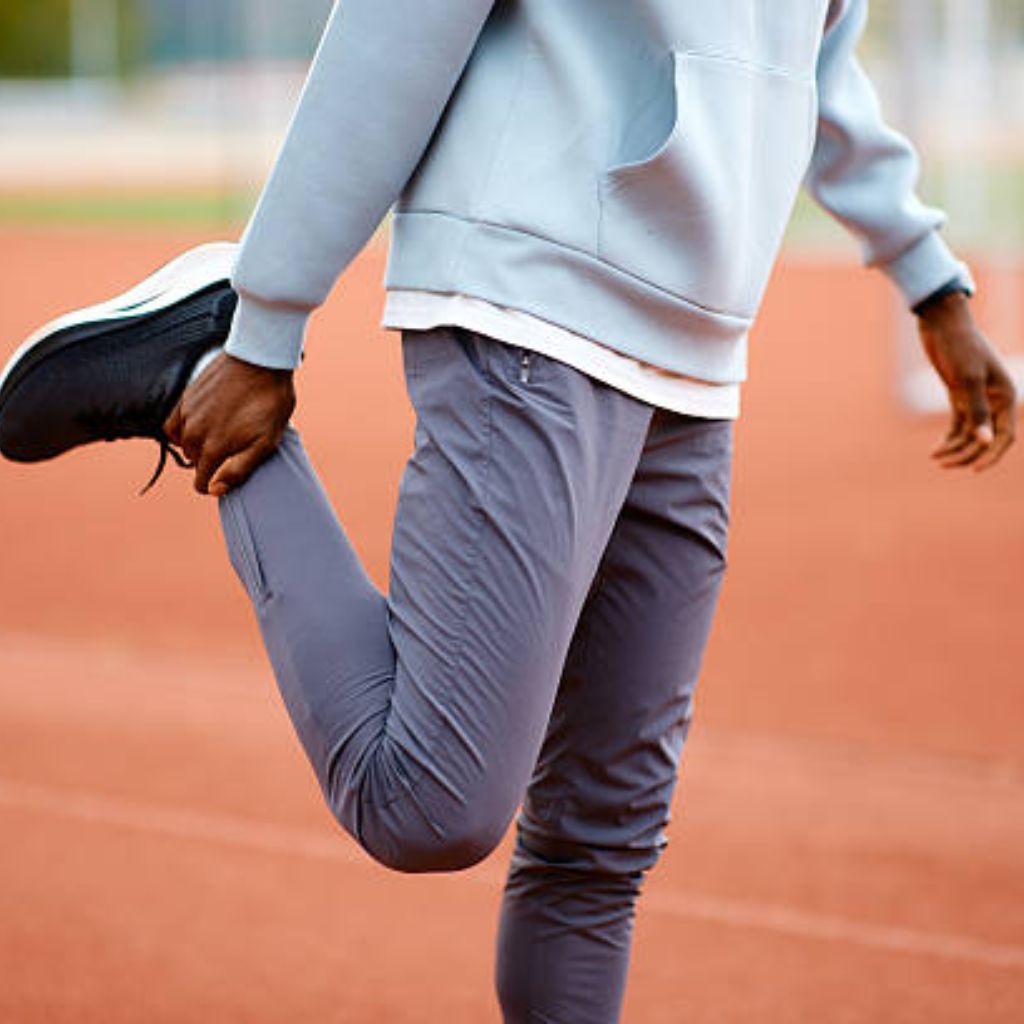 Kinetic Flow Athletic Fabric: Ultimate Comfort for Athletes 7 A person in a light blue hoodie and gray pants stretches their leg on a red athletic track, holding their foot with one hand.
Kinetic Flow Athletic Fabric: