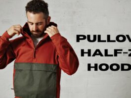 A man wearing a two-toned pullover hoodie (rust-colored top half and olive green bottom half) adjusts the hood while standing against an off-white, slightly textured wall. Large black text to the right reads “PULLOVER HALF-ZIP HOODIE.”