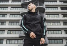 Techwear Half-Zip Hoodie: Trendy Urban Streetwear Guide A man wearing a black techwear tracksuit with turquoise stripes stands outdoors in front of a modern office building with a cylindrical staircase feature.