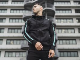A man wearing a black techwear tracksuit with turquoise stripes stands outdoors in front of a modern office building with a cylindrical staircase feature.