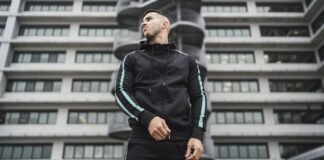 Techwear Half-Zip Hoodie: Trendy Urban Streetwear Guide A man wearing a black techwear tracksuit with turquoise stripes stands outdoors in front of a modern office building with a cylindrical staircase feature.