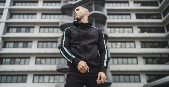 A man wearing a black techwear tracksuit with turquoise stripes stands outdoors in front of a modern office building with a cylindrical staircase feature.