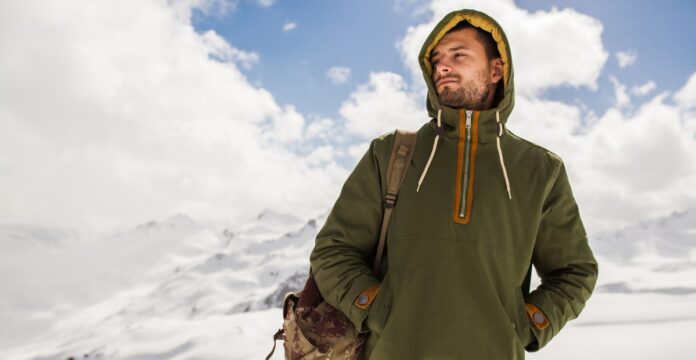 A man standing in a snowy mountain landscape, wearing an olive green hooded half-zip pullover with a tan zipper lining and drawstrings. He has a camouflage backpack over his left shoulder and is looking to the right, with snow-capped mountains and a partly cloudy sky in the background.