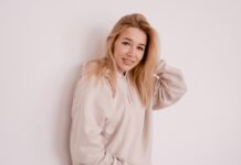 Vintage Half-Zip Hoodie: Classic Style & Daily Comfort Tips A young woman with long blonde hair wearing a light beige hoodie stands against a plain white wall, smiling softly with one hand touching her hair.