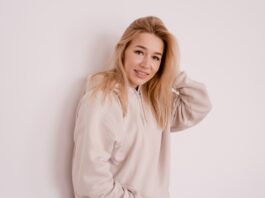 A young woman with long blonde hair wearing a light beige hoodie stands against a plain white wall, smiling softly with one hand touching her hair.