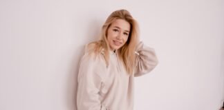 Vintage Half-Zip Hoodie: Classic Style & Daily Comfort Tips A young woman with long blonde hair wearing a light beige hoodie stands against a plain white wall, smiling softly with one hand touching her hair.
