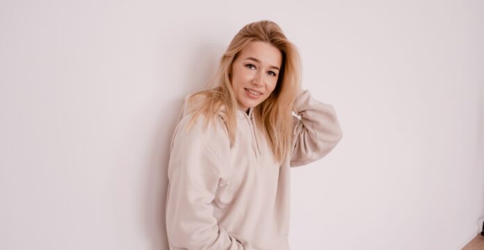 A young woman with long blonde hair wearing a light beige hoodie stands against a plain white wall, smiling softly with one hand touching her hair.