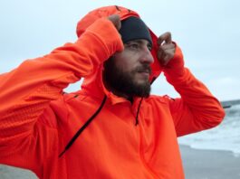 A bearded man on a beach wearing an orange hooded waterproof half-zip jacket, pulling the hood over his head while looking to the side; gray beanie on his head and an overcast sea background.