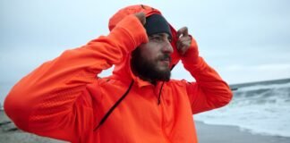 Waterproof Half-Zip Hoodie: Ultimate Guide to Style & Comfort A bearded man on a beach wearing an orange hooded waterproof half-zip jacket, pulling the hood over his head while looking to the side; gray beanie on his head and an overcast sea background.