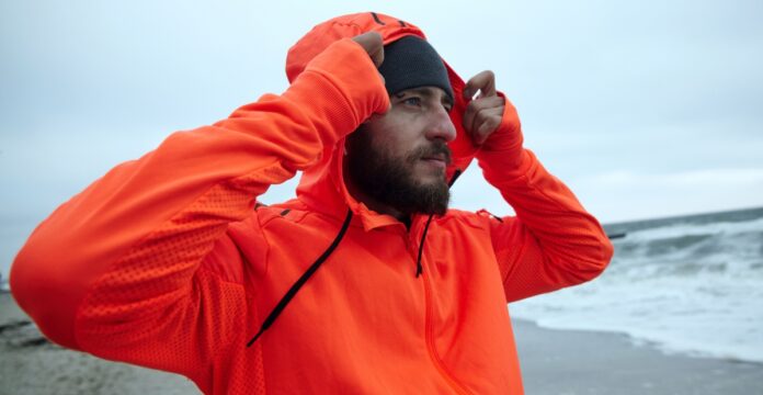 A bearded man on a beach wearing an orange hooded waterproof half-zip jacket, pulling the hood over his head while looking to the side; gray beanie on his head and an overcast sea background.