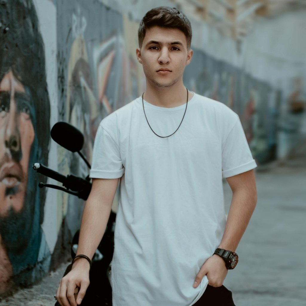 Model wearing a white crew neck t-shirt with a necklace and smartwatch, standing outdoors near a wall and a bicycle.

