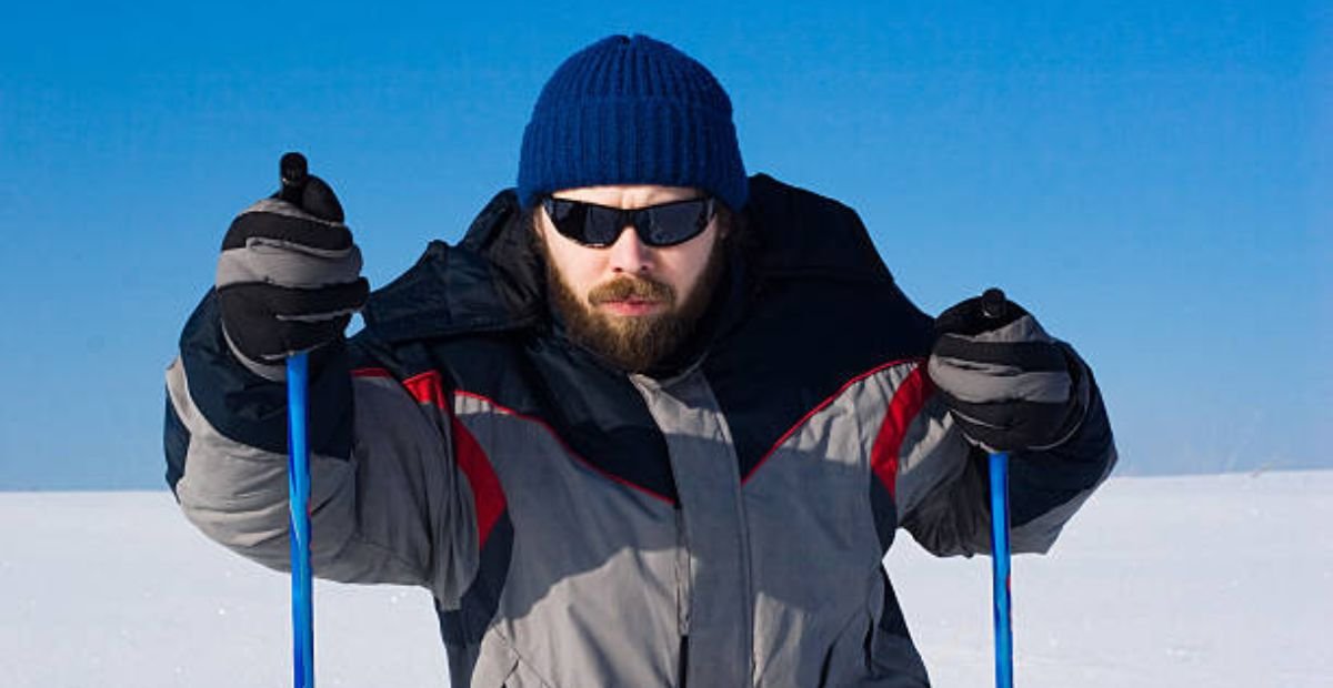 Arctic Pulse Sports Fabric: Engineered for Peak Performance "A bearded man wearing a blue beanie, sunglasses, and a high-performance winter coat stands confidently in deep snow, holding blue ski poles. Clear blue sky and expansive snowy landscape in background." Arctic Pulse Sports Fabric: