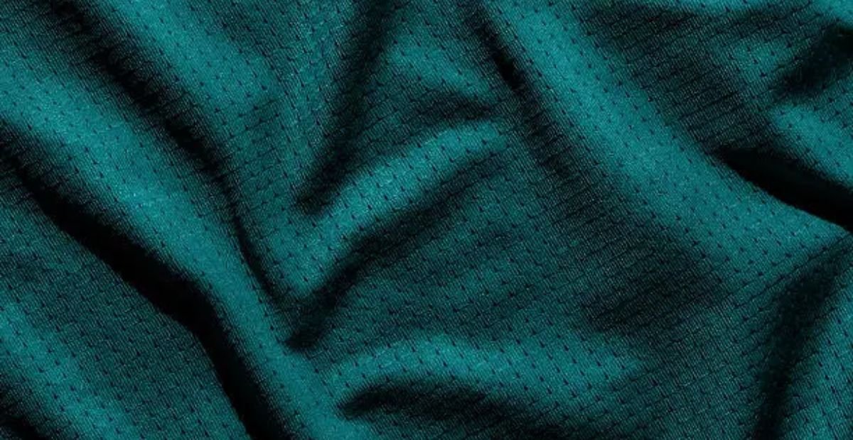 Turbo Dry Athletic Fabric: Stay Fresh, Dry & Focused All Day Close-up of a teal synthetic winter coat fabric with a subtle grid-pattern of perforations, shown draped with visible texture, shadows, and highlights. Turbo Dry Athletic Fabric: